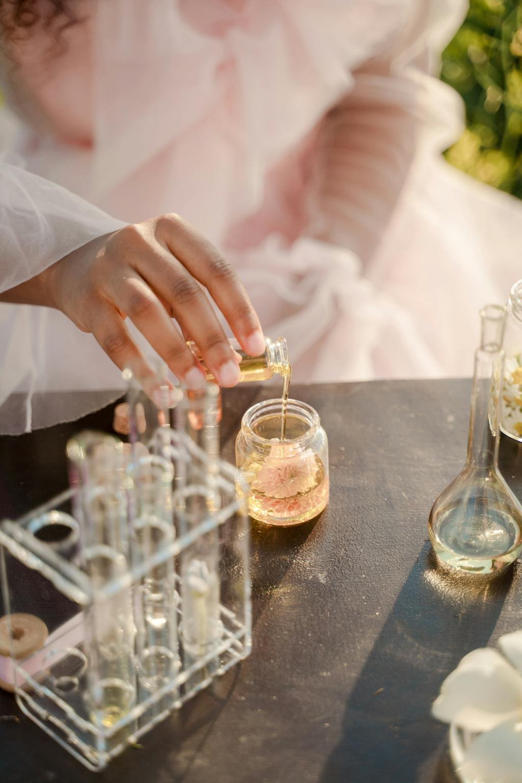 A woman in a pink dress pours liquid into a jar outdoors, surrounded by test tubes.